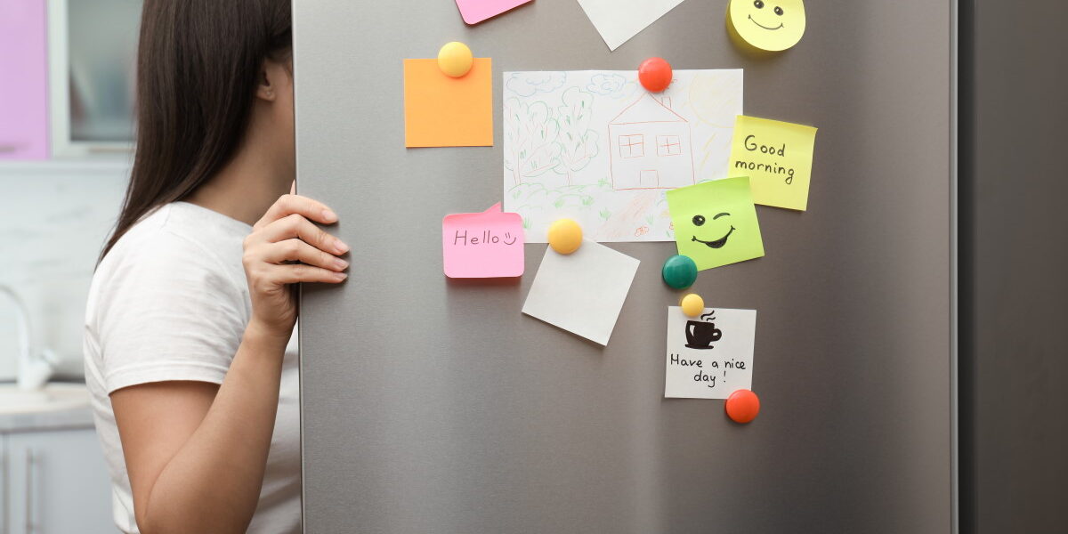 Woman opening refrigerator door with paper sheets and magnets at home, closeup